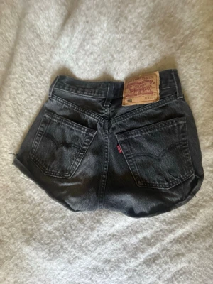 Svarta Levi's 501 jeansshorts - Säljer ett par svarta Levi's 501 jeansshorts med klassisk femficksdesign och uppvikta benslut. Shortsen har hög midja och den ikoniska läderlappen bak. Perfekta för sommaren och riktigt snygga till sneakers eller boots.