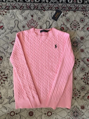 Rosa kabelstickad tröja Polo Ralph Lauren - Kabelstickad rosa tröja från Polo Ralph Lauren med rund halsringning och ribbade muddar. Tröjan har den klassiska broderade loggan i marinblått på bröstet och är tillverkad i mjukt material. Perfekt för en stilren och avslappnad look.
