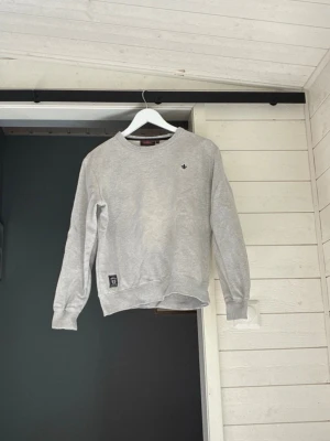 Morris sweatshirt  - En stilren ljusgrå sweatshirt med rund halsringning och svart broderad lilja på bröstet. Tröjan har ribbade muddar vid ärmslut och nederkant samt en liten patch nertill. Perfekt för en avslappnad look.