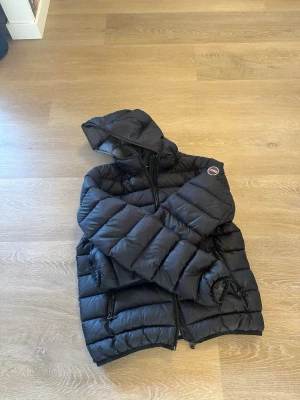 Svart dunjacka från Colmar - Snygg svart dunjacka från Colmar med huva och två dragkedjeförsedda fickor. Jackan har quiltad design och Colmar-logga på ärmen. Perfekt för kalla vinterdagar och riktigt skön att bära. Klassisk modell som funkar till det mesta.