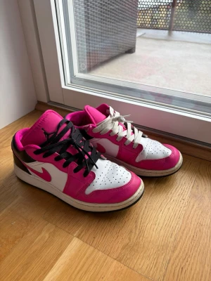 Nike Air Jordan 1 Low rosa och vita - Säljer ett par Nike Air Jordan 1 Low sneakers i vitt, rosa och svart. Skorna har klassisk låg siluett, perforerad tå och kontrasterande snörning i svart och vitt. Ikonisk Swoosh-logga på sidorna och mjukt lädermaterial. Perfekta för dig som vill sticka ut med färg.