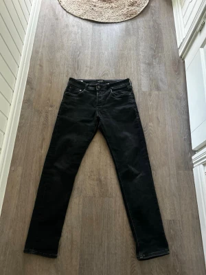 Svarta slim fit jeans Jack & Jones - Snygga svarta slim fit jeans från Jack & Jones med klassisk femficksmodell och knappgylf. Jeansen har en stilren look och passar perfekt till en avslappnad eller trendig outfit. Materialet är mjukt och stretchigt för extra komfort.