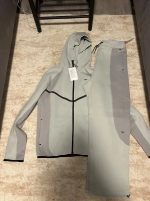 Nike grå träningsset med huva - Snyggt träningsset från Nike i ljusgrått med mörkgrå detaljer. Setet består av en hoodie med dragkedja och svarta linjer samt matchande byxor med dragsko i midjan. Perfekt för gymmet eller chill. Materialet är mjukt och stretchigt för maximal komfort.