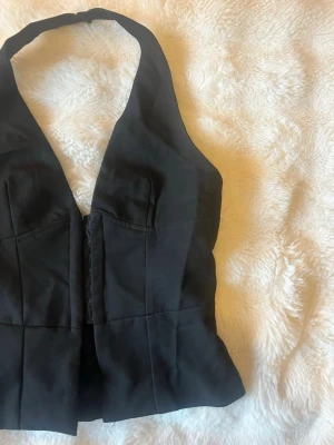 Svart halterneck väst med knäppning - Snygg svart väst med halterneck och djup v-ringning. Västen har markerade sömmar och knäppning framtill. Perfekt att styla över en skjorta eller t-shirt för en trendig look.