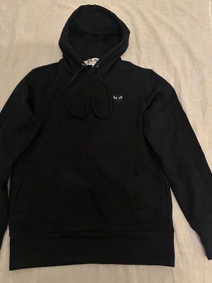 Play hoodie - Säljer en svart hoodie med huva och dragsnören. På bröstet finns en liten svart hjärtdetalj med ögon. Hoodien har fickor på sidorna och är tillverkad i mjukt material, perfekt för en chill och stilren look.