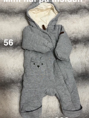 Grå overall med björndetalj, stl 56 - Mysig grå overall från newbie i storlek 56. Overallen har huva med vit insida och en söt ficka med björnansikte framtill. Tillverkad i mjukt material och har vikbara muddar vid ärmar och ben. Perfekt för kyliga dagar.🌸 Mini hål på insidan. 56