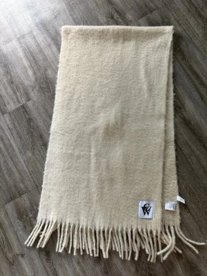 Beige halsduk från Carin Wester  - Mysig beige halsduk från Carin Wester i samarbete med Wilma. Halsduken är i mjuk, fluffig kvalitet med långa fransar och en stor vit patch med svart GW-logga. Perfekt för höst och vinter! Helt ny, i mycket fint skick. 