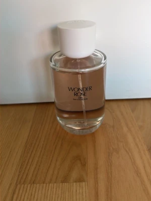 Wonder Rose parfym med vit kork - En elegant parfymflaska med ljusrosa doftvätska och vit rund kork. Flaskan har texten 'Wonder Rose' och en stilren, genomskinlig design som känns modern och fräsch. Perfekt för dig som gillar blommiga och fräscha dofter.