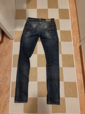 Nudie jeans - Säljer ett par mörkblå slim fit jeans med klassisk femficksdesign och snygga slitningar på framsidan. Jeansen har kontrastsömmar och raka ben. Perfekta till vardagslooken och passar till det mesta.