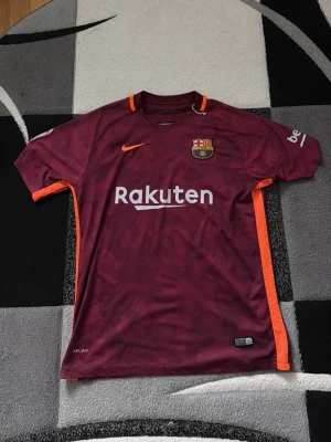 Barcelona Neymar Jr matchtröja vinröd - Säljer en vinröd FC Barcelona matchtröja från Nike med Neymar Jr och nummer 11 på ryggen. Tröjan har orange detaljer längs sidorna, klubbmärke på bröstet och Rakuten som sponsor framtill. Tillverkad i Dri-FIT material för bästa komfort.