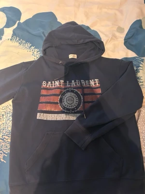Mörkblå hoodie med Saint Laurent-tryck - Snygg mörkblå hoodie med huva och känguruficka. Framsidan har ett stort Saint Laurent-tryck i vitt och rött med slitna detaljer och emblem. Perfekt för dig som gillar streetwear och vill ha en bekväm och stilren look.