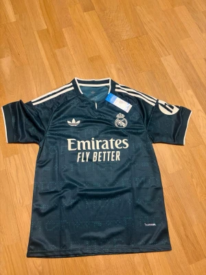 BELLINGHAM 26/27 borta S - Snygg mörkblå Real Madrid matchtröja från Adidas med Bellingham och nummer 5 på ryggen. Tröjan har vita detaljer, klubbmärke och sponsortryck framtill samt klassiska Adidas-ränder på axlarna. Tillverkad i lätt och ventilerande material.