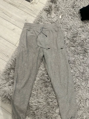 Grå mjukisbyxor  - Snygga grå mjukisbyxor från Lacoste med dragsko i midjan och resår vid bensluten. Byxorna har sidofickor och den klassiska Lacoste-loggan broderad på ena benet. Perfekta för en chill och avslappnad stil. Står Xl men passar S