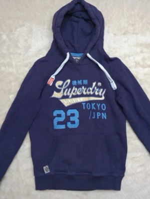 Mörkblå Superdry hoodie med tryck - Snygg mörkblå hoodie från Superdry med stort tryck på bröstet i vitt, blått och gult. Hoodien har huva med vita dragsnören och känguruficka framtill. Perfekt för en avslappnad streetwear-look. Lite smutsig men syns knappast 