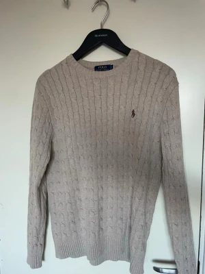 Tröja Polo Ralph Lauren - Kabelstickad tröja i beige från Polo Ralph Lauren med rund halsringning och den klassiska broderade loggan på bröstet. Långa ärmar och ribbade muddar vid ärmslut och nederkant. Perfekt för lager på lager och en stilren look.