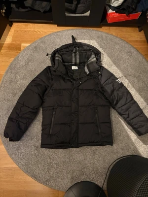 Pufferjacka Burberry London England - Helt ny Snygg svart pufferjacka med huva och dragkedja samt knappar framtill. Jackan har två sidofickor med dragkedja och ett rutigt innerfoder i grått och svart. Perfekt för kalla vinterdagar och har en modern, avslappnad passform. Orginal påse följer med när jag köpte jackan 