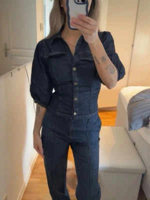 Mörkblå denim blus med puffärm & byxor  - Säljer en mörkblå denimblus med markerad midja, puffiga trekvartsärmar och knappar framtill. Blusen har två bröstfickor och tydliga sömmar som ger en cool vintagekänsla. Perfekt att styla med jeans för en matchande look. Säljer även byxorna om man vill ha de som ett set. Byxorna är i storlek 36 och tröjan S