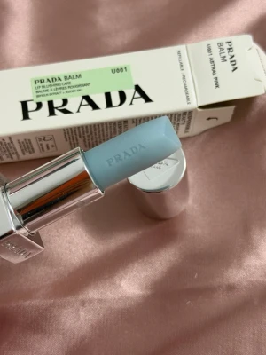 Prada Balm i ljusblå nyans - Lyxigt läppbalsam från Prada i en snygg ljusblå färg. Kommer i en elegant silverfärgad hylsa med Pradas logga ingraverad på både balsamet och hylsan. Förpackningen är stilren och modern, perfekt för dig som gillar exklusiva detaljer.