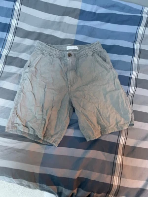 Gråa bomullsshorts med fickor - Säljer ett par gråa shorts i bomull med normal passform. Shortsen har sidofickor, knapp och dragkedja framtill samt bälteshällor i midjan. Perfekta för varma dagar och enkel stil.