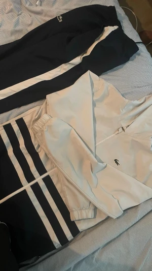 Blåvit tracksuit  Lacoste(nypris 3000kr) - Snygg blåvit tracksuit från Lacoste med dragkedja på jackan och elastiska muddar. Jackan har två mörkblå ränder på nedre delen och byxorna har vit sidoremsa. Perfekt för sport eller chill. Klassisk krokodillogo på både jacka och byxor. Vill få bort så snabbt så möjligt , billigare vid snabb affär, passar som S också , fraktar direkt , om du är intresserad eller har några frågor , eller ha mer bilder så är det bara att höra av sig.