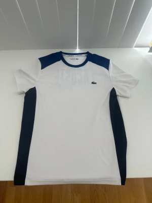 Vit och blå Lacoste t-shirt äkta sällsynt!! - Säljer en vit t-shirt från Lacoste med blå detaljer på axlar och sidor. T-shirten har grönt Lacoste-tryck och krokodil på ryggen samt liten krokodillogo fram. Klassisk rund hals och korta ärmar. Perfekt för en sportig och clean look. Sällsynt omöjligt hitta idag! Äkta Lacoste 
