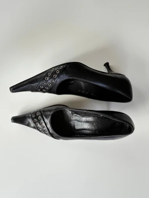 Svarta pumps med nitar och spetsig tå - Säljer ett par svarta pumps i skinn med spetsig tå och dekorativa silverfärgade nitar längs sidan. Klacken är smal och elegant, och skorna har en stilren design som sticker ut.
