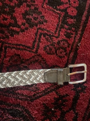 Flätat beige och brunt bälte - Snyggt flätat bälte i beige och bruna toner med silverfärgat spänne. Bältet har detaljer i brunt skinn vid spännet och passar perfekt till jeans eller chinos. Enkel att justera tack vare det flexibla flätade materialet. 100cm långt