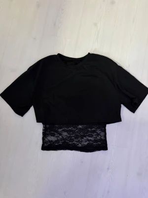 Svart croppad topp med spetsdetalj - Snygg svart croppad t-shirt med korta ärmar och en unik spetsdetalj nedtill. Toppen har en loose passform och rund halsringning. Perfekt för dig som vill ha en trendig och edgy look. Köpt från newyorker men lappen är bortklippt