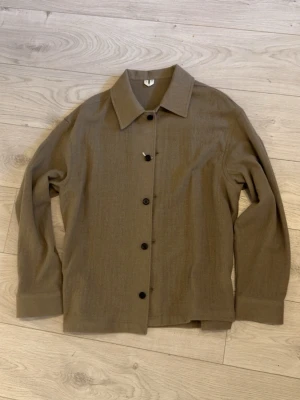 Beige Arket overshirt  - Stilren beige overshirt med klassisk krage och svarta knappar framtill. Overshirten har långa ärmar och är tillverkad i ett mjukt, vävt material som ger en avslappnad look. Perfekt att bära över en t-shirt för en enkel och trendig stil. Storlek: M