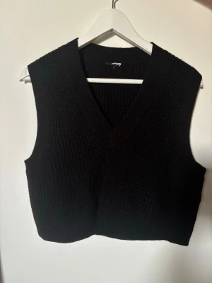 Svart stickad väst med v-ringning - Säljer en svart stickad väst med bred v-ringning och ribbad struktur. Västen är croppad och har en klassisk, enkel design som passar till många olika outfits. Perfekt att bära över en skjorta eller t-shirt för en trendig look.