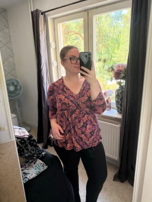 Blommig lila blus med v-ringning - Supersöt blus i lila med blommigt mönster i orange och rosa toner. Blusen har v-ringning, knytband vid halsen och långa ärmar. Modellen är lite lösare och ger en avslappnad känsla. Perfekt att styla med jeans eller kjol för en trendig look. Från märket Odd Molly i storlek 3 som enligt deras storlekguide motsvarar en storlek L. Jag har vanligtvis en 4/XL. Fin men den ör blekare än ny. Kan vara något tvätt påverkad. 