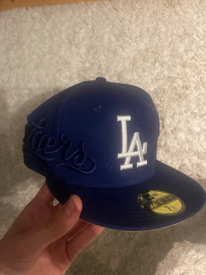 Blå LA Dodgers keps från New Era - Snygg blå snapback-keps från New Era med LA Dodgers-logga broderad i vitt framtill och Dodgers-text på sidan. Klassisk 59FIFTY-modell med platt skärm och broderade detaljer. Perfekt för dig som gillar streetwear och sportig stil.