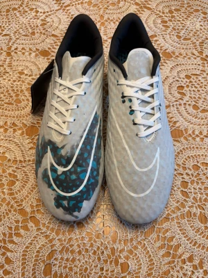 Nike fotbollsskor  - Snygga vita fotbollsskor från Nike med stora vita swoosh-loggor på båda sidor. Ena skon har ett blått och svart kamouflagemönster på sidan, medan den andra är helvit. Skorna har vita snören och svart insida. Perfekta för dig som vill sticka ut på planen.
