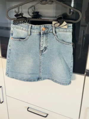 Ljusblå jeanskjol med fickor - Snygg ljusblå jeanskjol i klassisk modell med fem fickor, bälteshällor och knappstängning fram. Kjolen är kort och har en rak passform. Enkel och tidlös denimstil som funkar till många outfits.