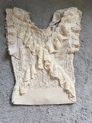 Beige spetsdetaljerad topp från Zara - Supersöt beige topp från Zara med virkade detaljer och volanger i spets. Toppen har v-ringning och är ärmlös med breda axelpartier. Perfekt för dig som gillar romantisk och bohemisk stil.
