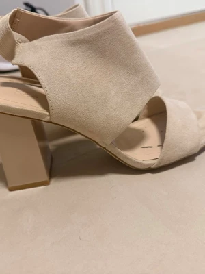Beige sandaler med bred klack - Säljer ett par stilrena beige sandaler med bred klack och öppen tå. Skorna har breda remmar i mockaimitation och en stabil fyrkantig klack som ger både höjd och komfort. Perfekta för dig som vill ha en enkel men trendig look.