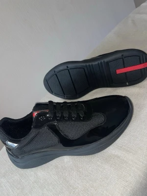 Svarta sneakers  - Snygga svarta sneakers med glansig finish och meshdetaljer. Skorna har svarta snören, diskret röd logga på plösen och sulan, samt en chunky sula för extra attityd. Perfekta för dig som vill ha en clean och modern look.