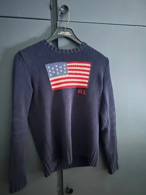 Mörkblå stickad tröja Polo Ralph Lauren - Mörkblå stickad tröja från Polo Ralph Lauren med amerikansk flagga i rött, vitt och blått på bröstet samt RL-broderi. Rund halsringning och ribbstickade muddar. Perfekt för dig som gillar klassisk preppy stil.