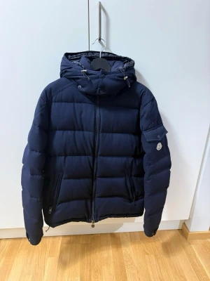 Mörkblå vinterjacka  från Moncler -  Mörkblå dunjacka från Moncler med huva och dragkedja framtill. Storlek 5 ( L)  har två sidofickor med dragkedja och en ficka med Moncler-logga på ärmen. Kontoutdrag finns på jackan, köpt från Kicks N Stuff i Göteborg. 