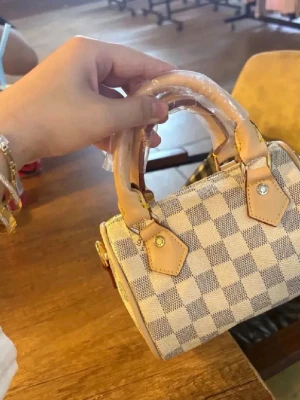 Rutig minihandväska i beige och vitt - Jag har köpt den på Plick men kommer inte till användning. En längre band medföljer även. Snygg minihandväska med rutigt mönster i beige och vitt. Väskan har två korta handtag i ljust läder och detaljer i guld. Perfekt accessoar för att lyfta din outfit med en trendig touch.