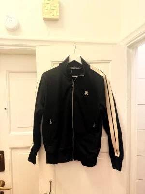 Palm Angels Monogram Track Jacket, Svart  -  Palm Angels Monogram Track Jacket i svart. Storlek M.  Knappt använd. Inget slitage, inga fläckar, inga hål. Dragkedjor och muddar är som nya. Off white-ränder längs ärmarna. Broderad PA-monogram på bröstet. Två dragkedjefickor fram.  Modell från 2024. Nypris cirka 7 000 kr.  Certilogo-tagg sitter kvar i plagget. Äktheten kan skannas och verifieras på plats.  Pris: 3 495 kr  Hämtas i Stockholm eller skickas mot frakt. Skriv i PM.