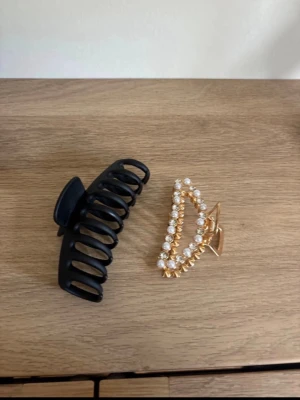 Två hårklämmor - Säljer två hårklämmor – en enkel svart i plast och en guldig med pärlor och glittrande stenar. Perfekta för att snabbt fixa frisyren och ge en snygg touch till håret.