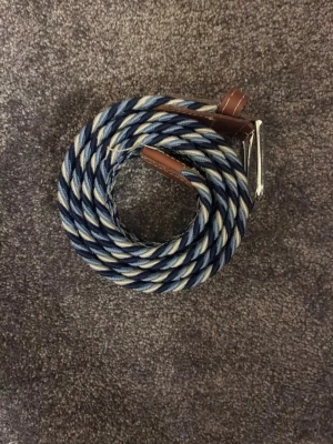 Blåvitt flätat tygbälte - Snyggt flätat bälte i blått och vitt tyg med bruna läderdetaljer vid spännet. Bältet har ett klassiskt silverfärgat metallspänne och passar perfekt till jeans eller chinos. Den flätade designen ger en avslappnad men stilren look.