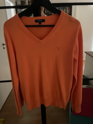  GANT - Snygg orange V-ringad tröja från GANT med diskret logga broderad på bröstet. Tröjan har långa ärmar och ribbade muddar vid ärmslut och nederkant. Perfekt för dig som vill ha en färgklick i garderoben.