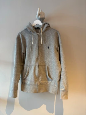 Grå zip hoodie från Polo Ralph Lauren - Snygg grå hoodie med dragkedja från Polo Ralph Lauren. Klassisk huva med vita snören och två fickor framtill. Liten mörkblå logga broderad på bröstet. Tillverkad i mjukt bommulsmaterial. 