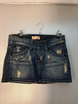 Minikjol denim - Snygg mörkblå jeanskjol i kort modell med slitna detaljer och klassiska fickor fram. Kjolen har bälteshällor och en knappstängning i midjan. Perfekt för dig som gillar en avslappnad och trendig stil.