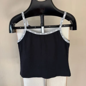 Svart linnetopp från Calvin Klein - Snygg svart linnetopp från Calvin Klein med tunna axelband och logotypdetaljer längs kanterna. 0380