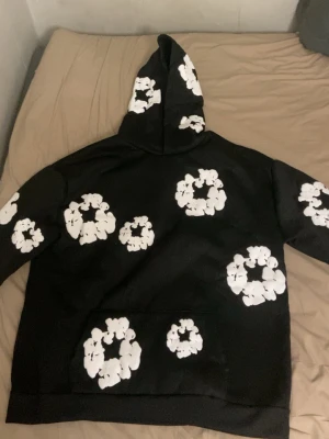 Svart hoodie med vita blommor - Säljer en svart hoodie med huva och stora vita blommönster över hela tröjan, även på huvan. Hoodien har en klassisk passform och magficka framtill. Perfekt för dig som vill sticka ut med en unik och trendig stil.