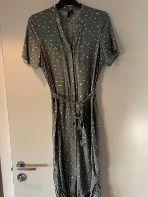 Grön mönstrad klänning Vero Moda - Snygg grön långklänning från Vero Moda med vitt blommönster. Klänningen har korta ärmar, v-ringning och ett matchande knytband i midjan. Tillverkad i ett mjukt och lätt material som passar perfekt till varmare dagar.