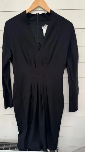 Max Mara Svart långklänning med v-ringning - Stilren svart långklänning från Max Mara med v-ringad hals och långa ärmar. Klänningen har markerad midja och veckade detaljer framtill som ger en snygg siluett. Dragkedja bak för enkel påklädning. Perfekt för dig som gillar klassisk och enkel stil.0121